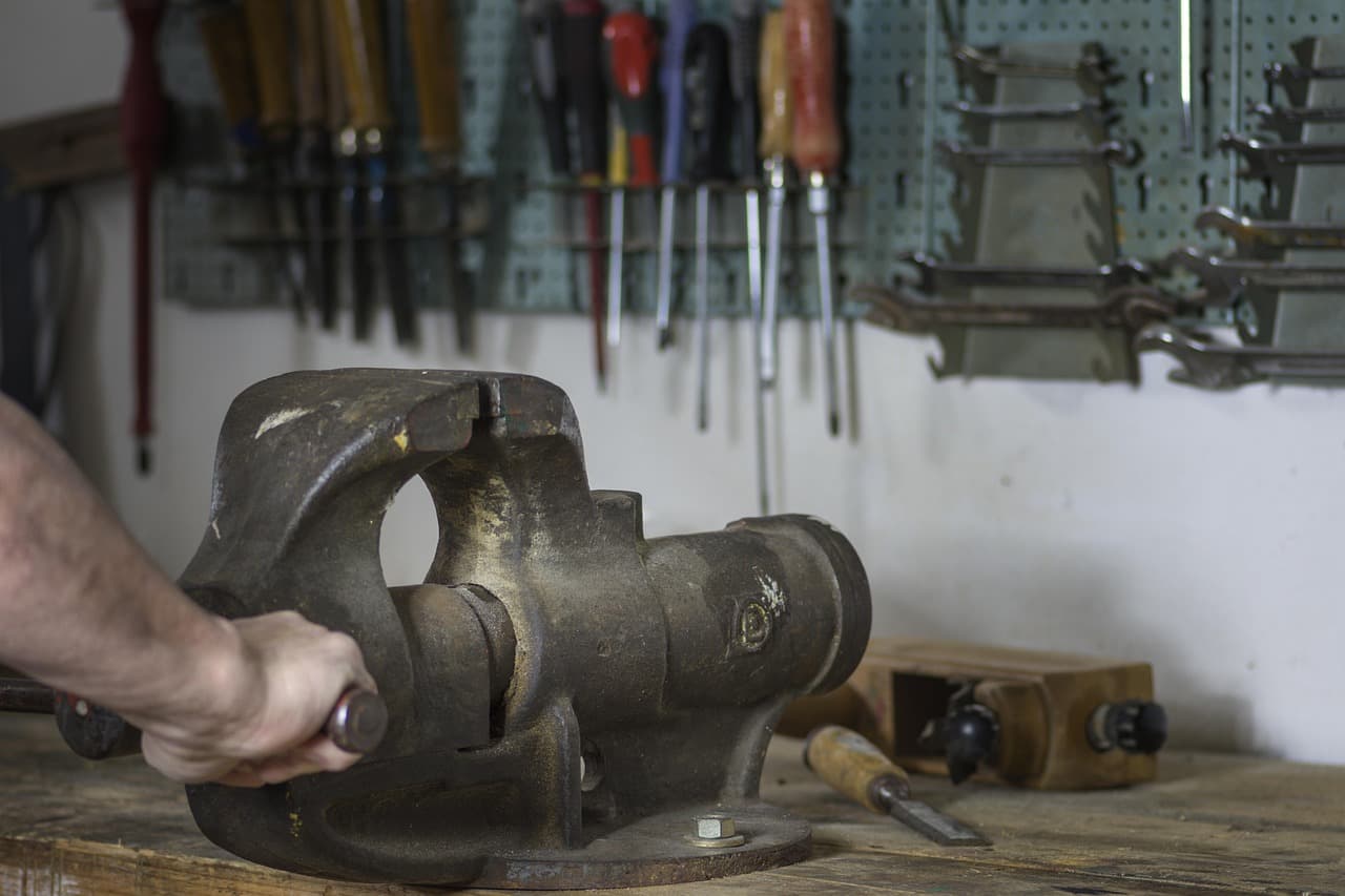 A hand working a bench vice in front of a wall of hand tools